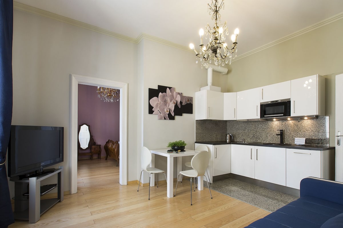 Een moderne woonkamer met een flatscreen televisie, een blauwe bank, een eethoek met een witte tafel en stoelen, en een keuken met witte kastjes en een magnetron.