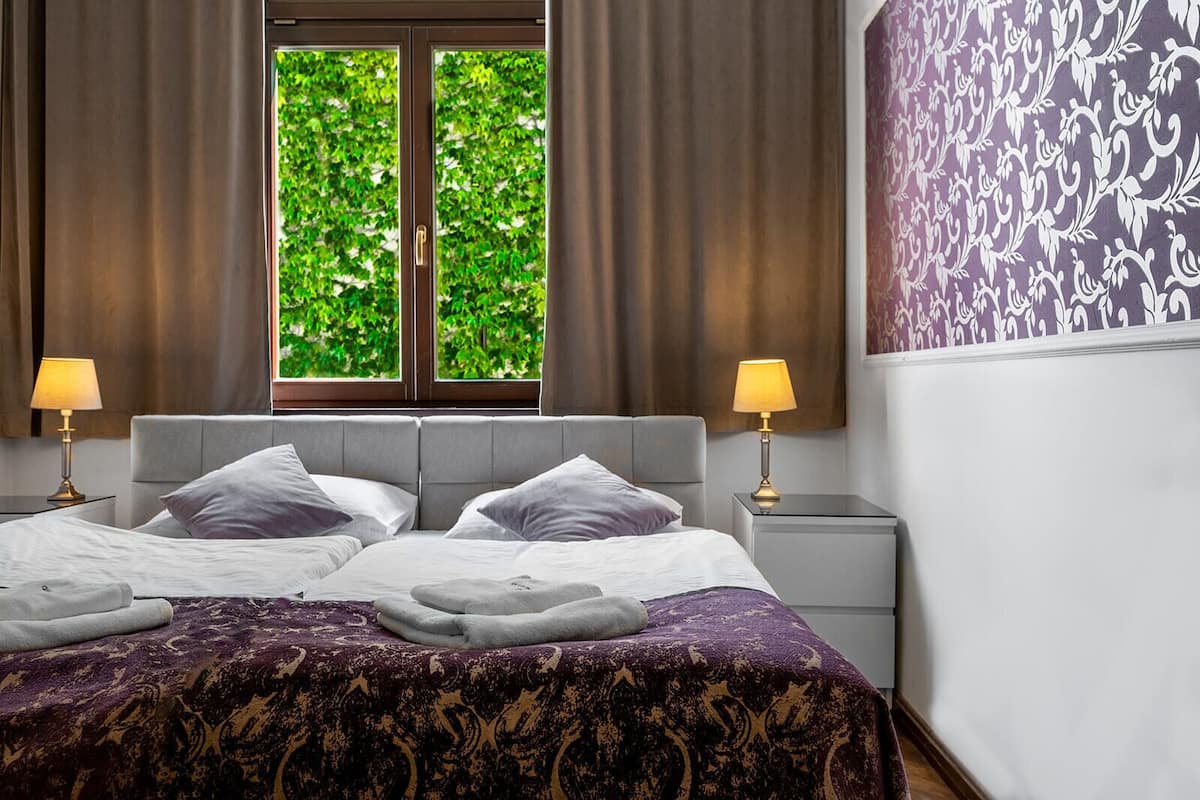 Een hotelkamer met een bed, twee bedlampen, een raam met uitzicht op groen en een behang met patroon.
