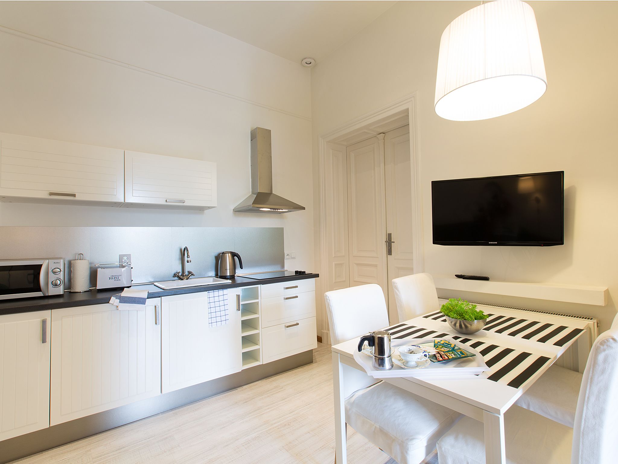 A modern kitchen with white cabinetry, a stainless steel range hood, a microwave, a kettle, and a toaster. There is a dining table with a black and white striped table runner, a white chair, and a small plant. A flat-screen TV is mounted on the wall above the dining area.