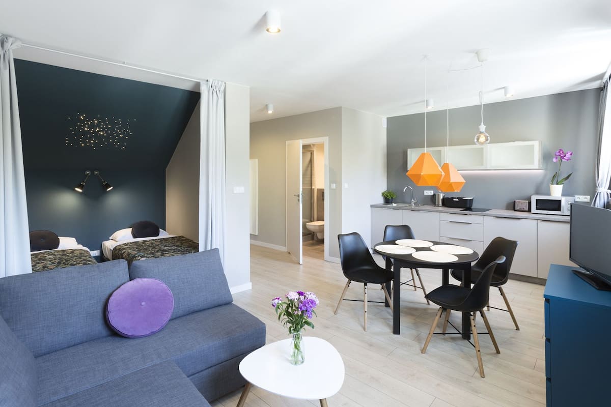 Een moderne woonkamer met een grijze bank, een klein wit tafeltje met bloemen, en een keuken met een eettafel en stoelen.