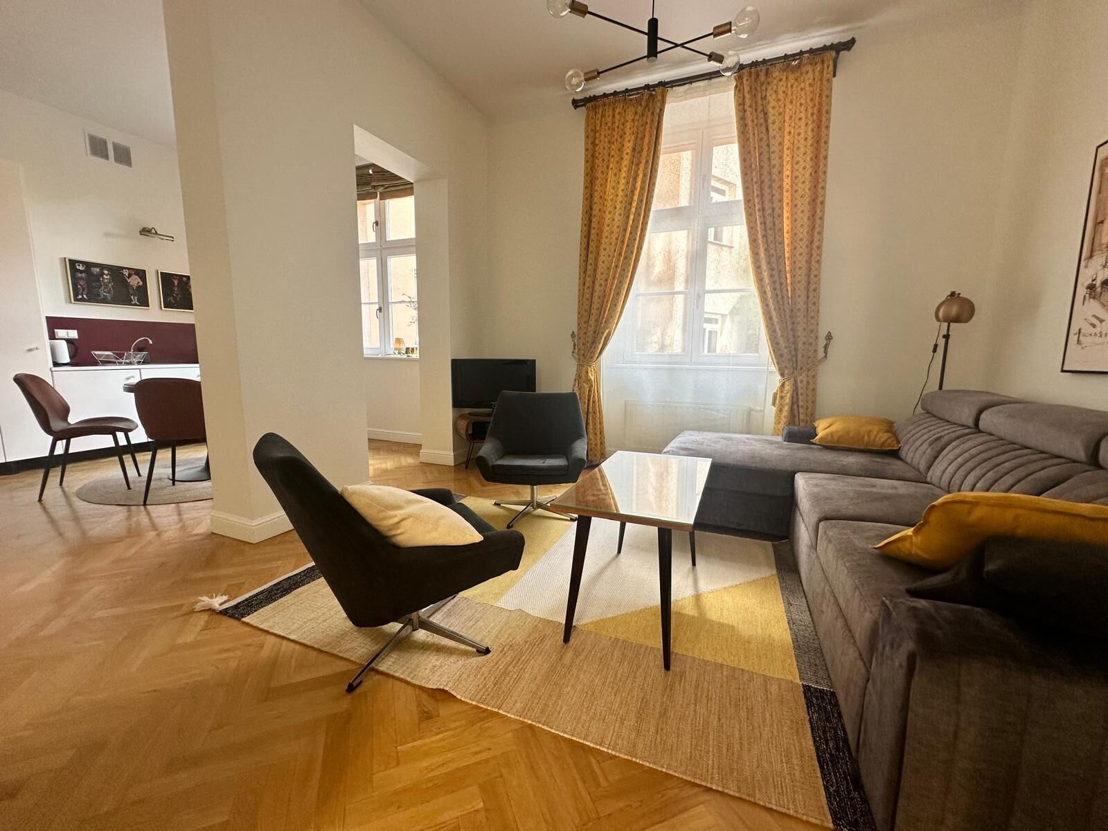 A modern living room with a sofa, armchair, and coffee table. There is a television and a dining area in the background.