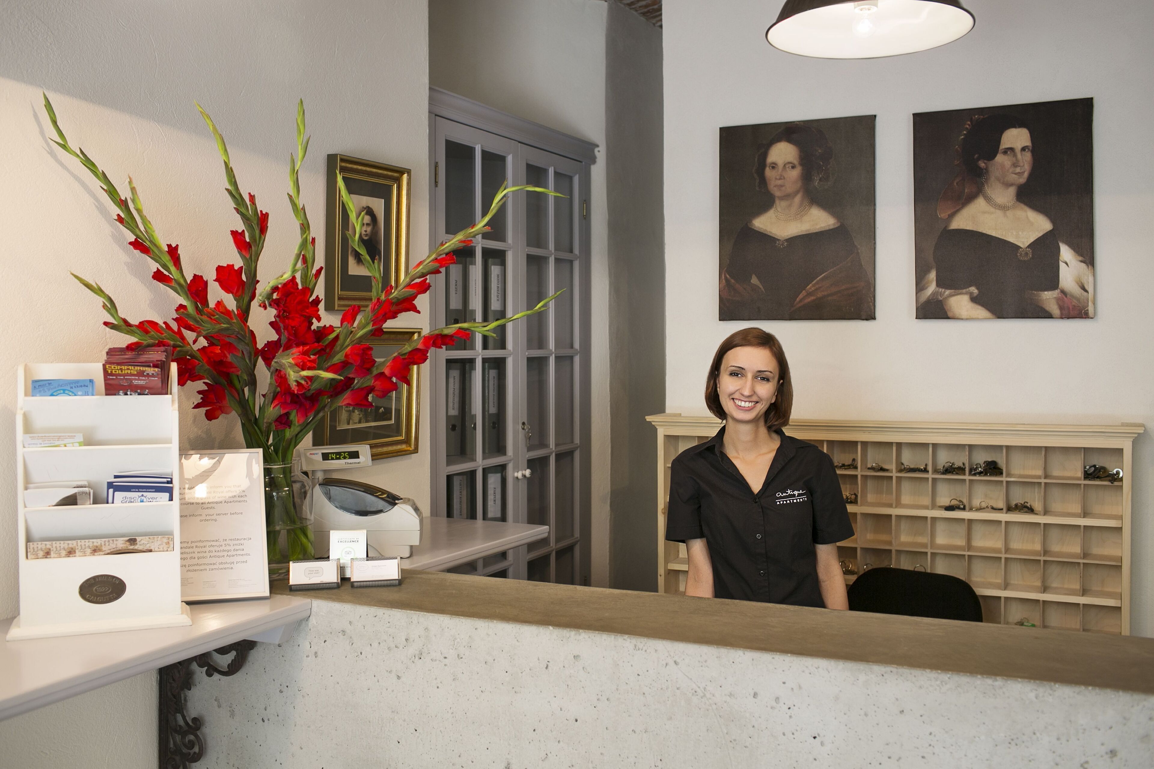 Un comptoir d’accueil orné d’un vase de fleurs rouges, des tableaux encadrés accrochés au mur, ainsi qu’un présentoir de livres et une horloge.