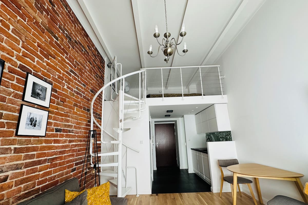 Een moderne loft met zichtbare bakstenen muren, een witte spiraaltrap, een eetgedeelte met een houten tafel en stoelen, en een kroonluchter.