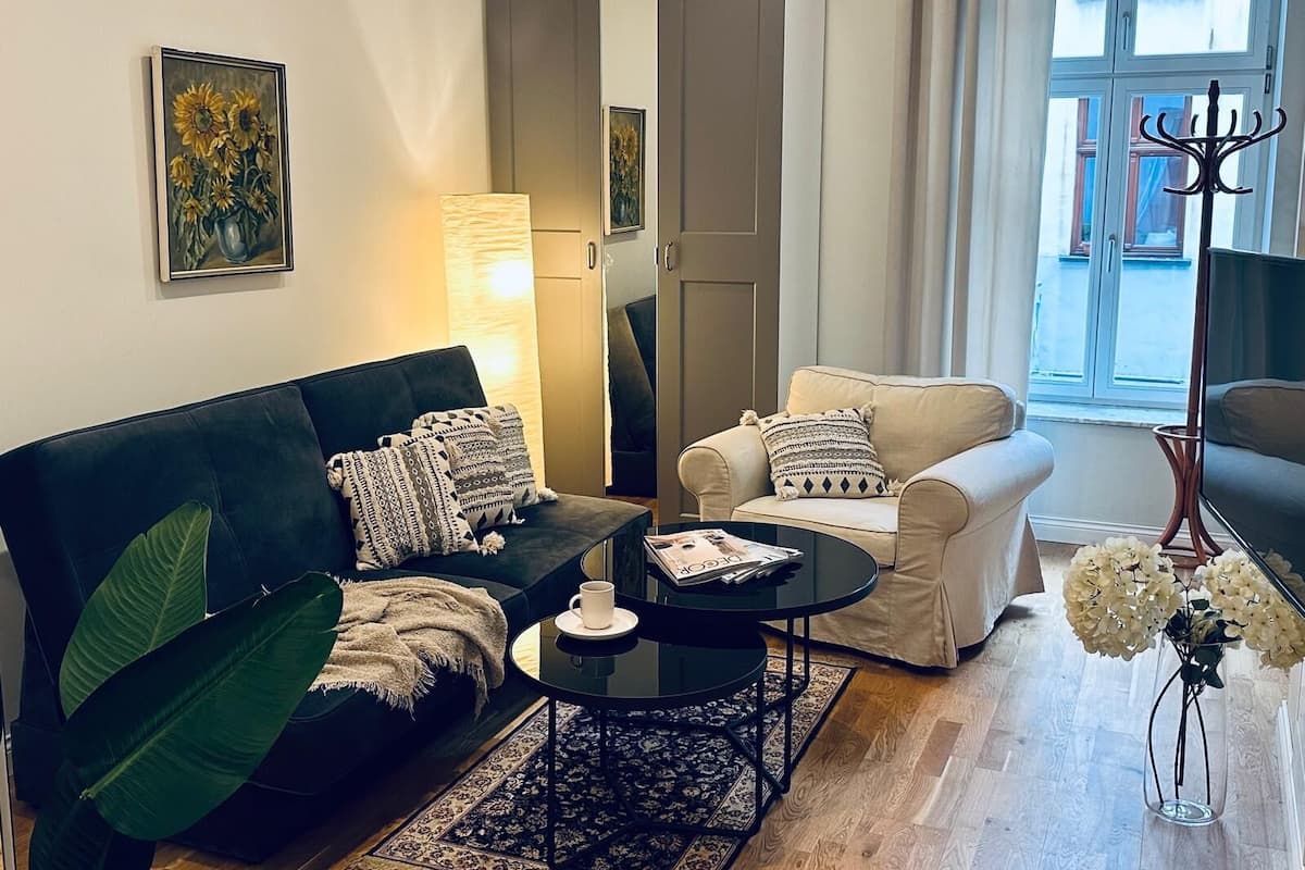 Een woonkamer met een zwarte bank, een beige bank, een salontafel en een vloerlamp. Er is een raam met gordijnen en uitzicht op gebouwen.