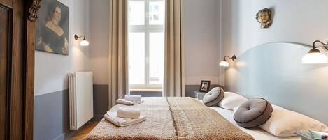 Ein ordentlich eingerichtetes Schlafzimmer mit einem großen Bett, einem Nachttisch, einem Heizkörper und einem Fenster mit Vorhängen.