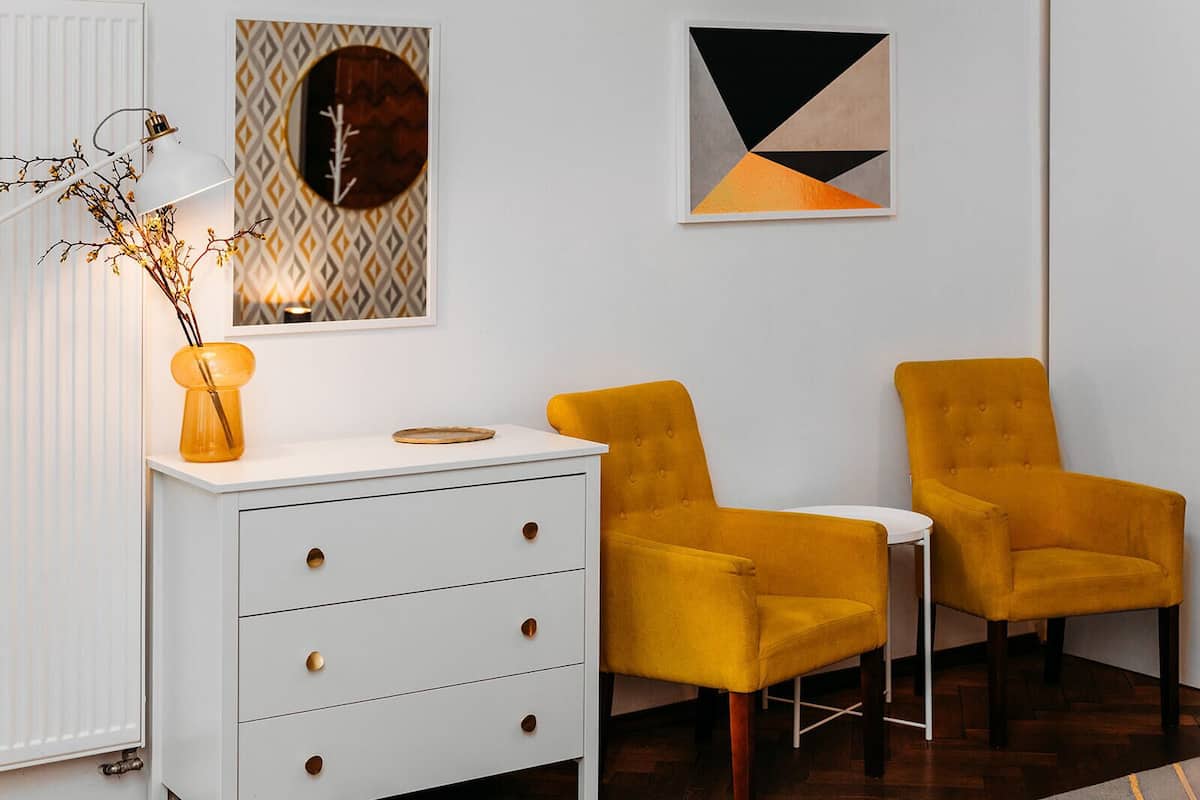 Een moderne kamer met een witte dressoir, twee mosterdkleurige fauteuils, een lamp en abstracte wanddecoratie.