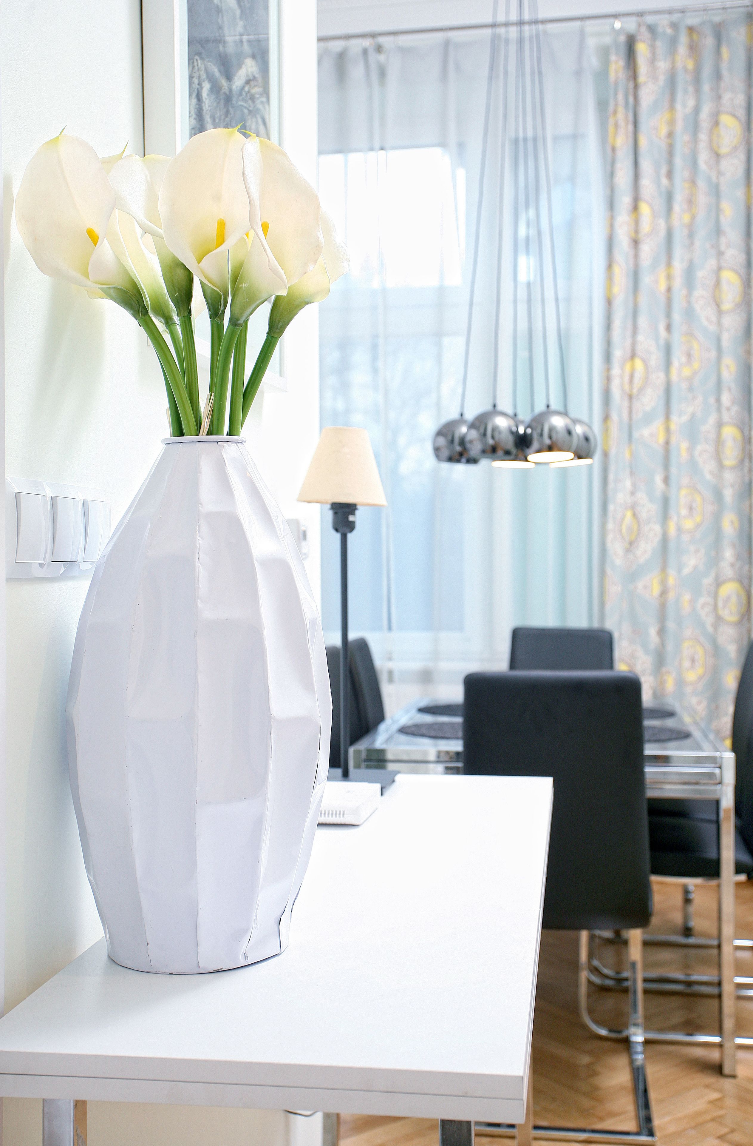 A white vase with calla lilies on a white table in a modern dining area with a chandelier and patterned curtains.
