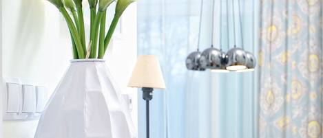 A white vase with calla lilies on a white table in a modern dining area with a chandelier and patterned curtains.