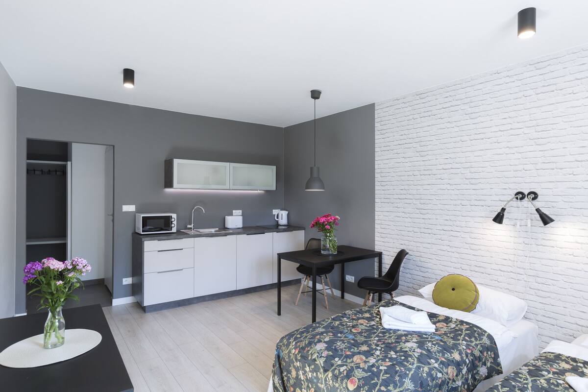 Een moderne kamer met een bed, een klein bureau, een stoel, een tafel met een bloevas en een kitchenette.