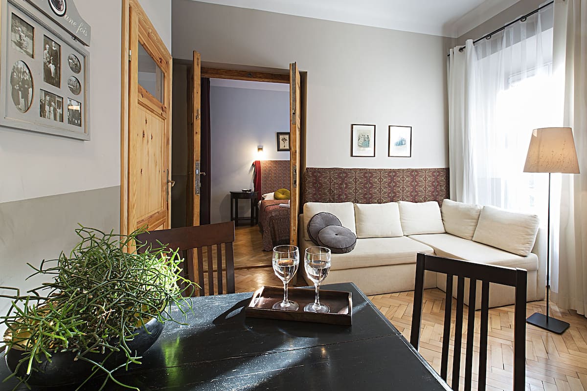 Een moderne woonkamer met een bank, een eettafel met een plant en wijnglazen, een houten deur en ingelijste foto's aan de muur.