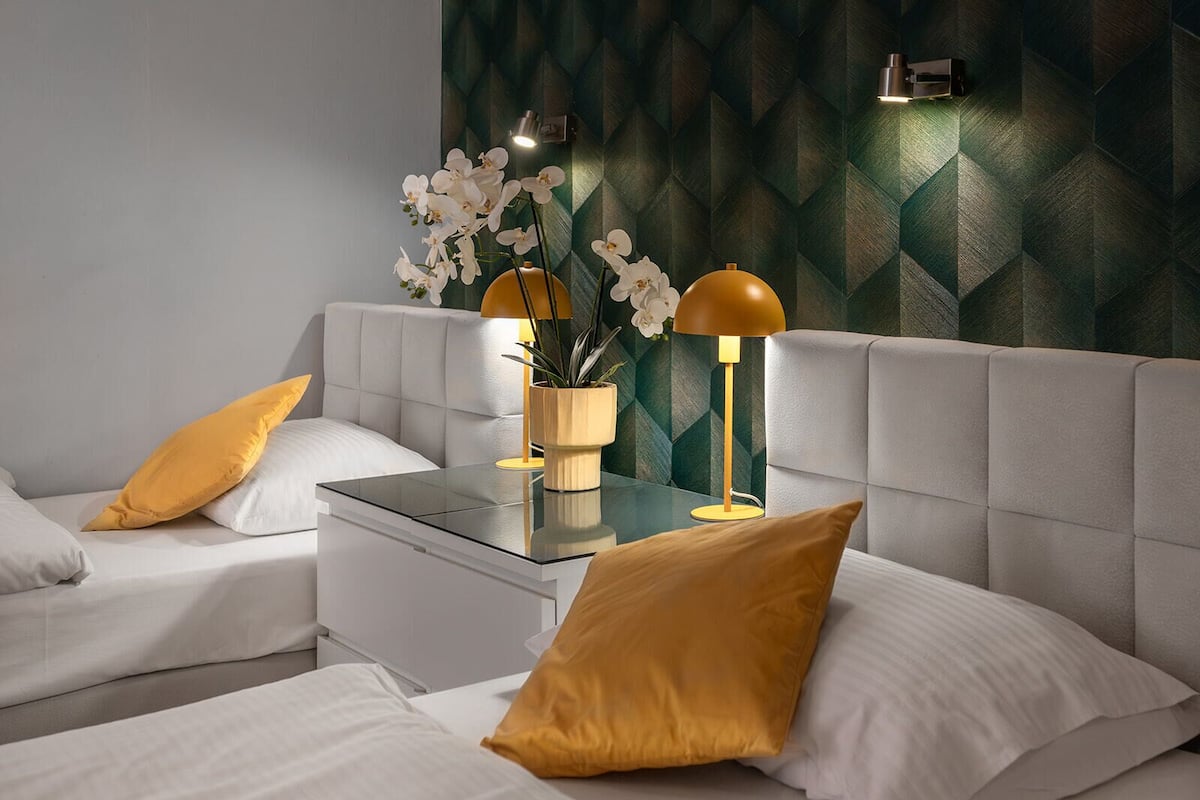 Een moderne slaapkamer met een gestoffeerd hoofdeinde, een glazen nachtkastje, twee witte bedden met gele kussens, een gele lamp en witte orchideeën in een houten vaas.