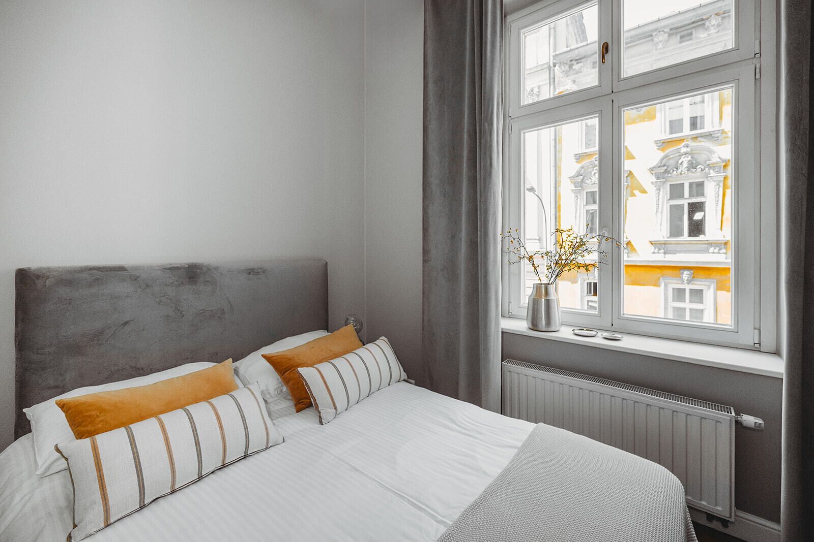 Une chambre à coucher avec un lit, des oreillers, une fenêtre donnant sur des immeubles et un radiateur.