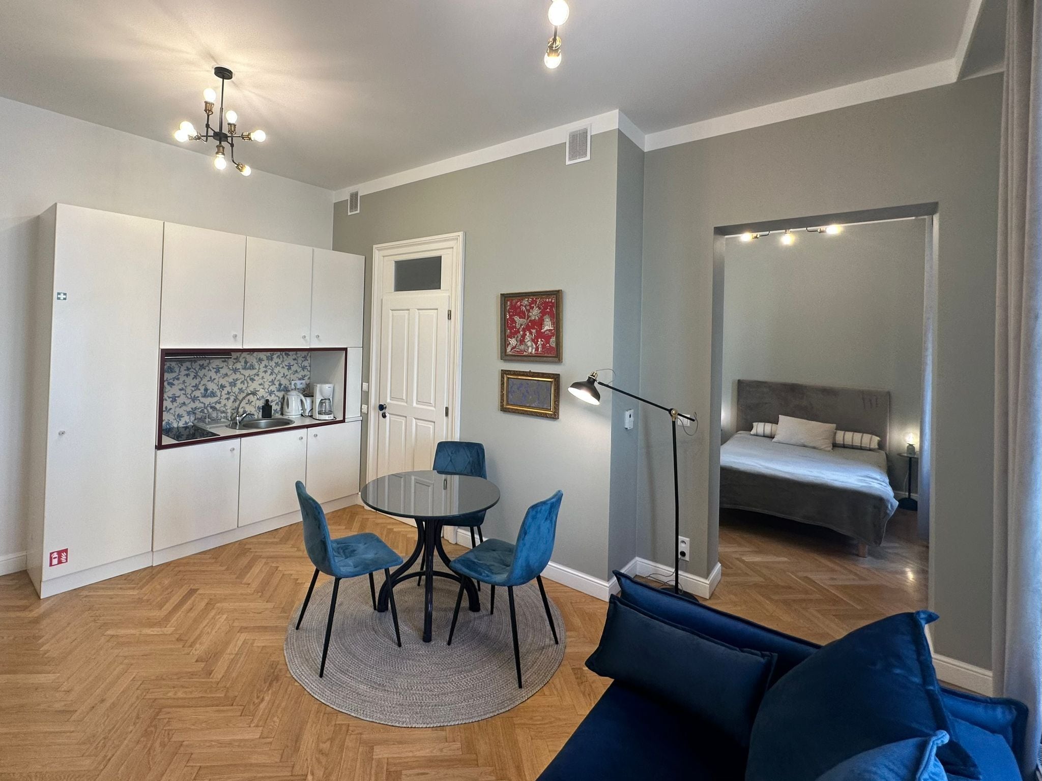 Ett kompakt bostadsutrymme med ett litet kök, en matplats med ett runt bord och stolar, en säng med en sänggavel och en stor spegel.