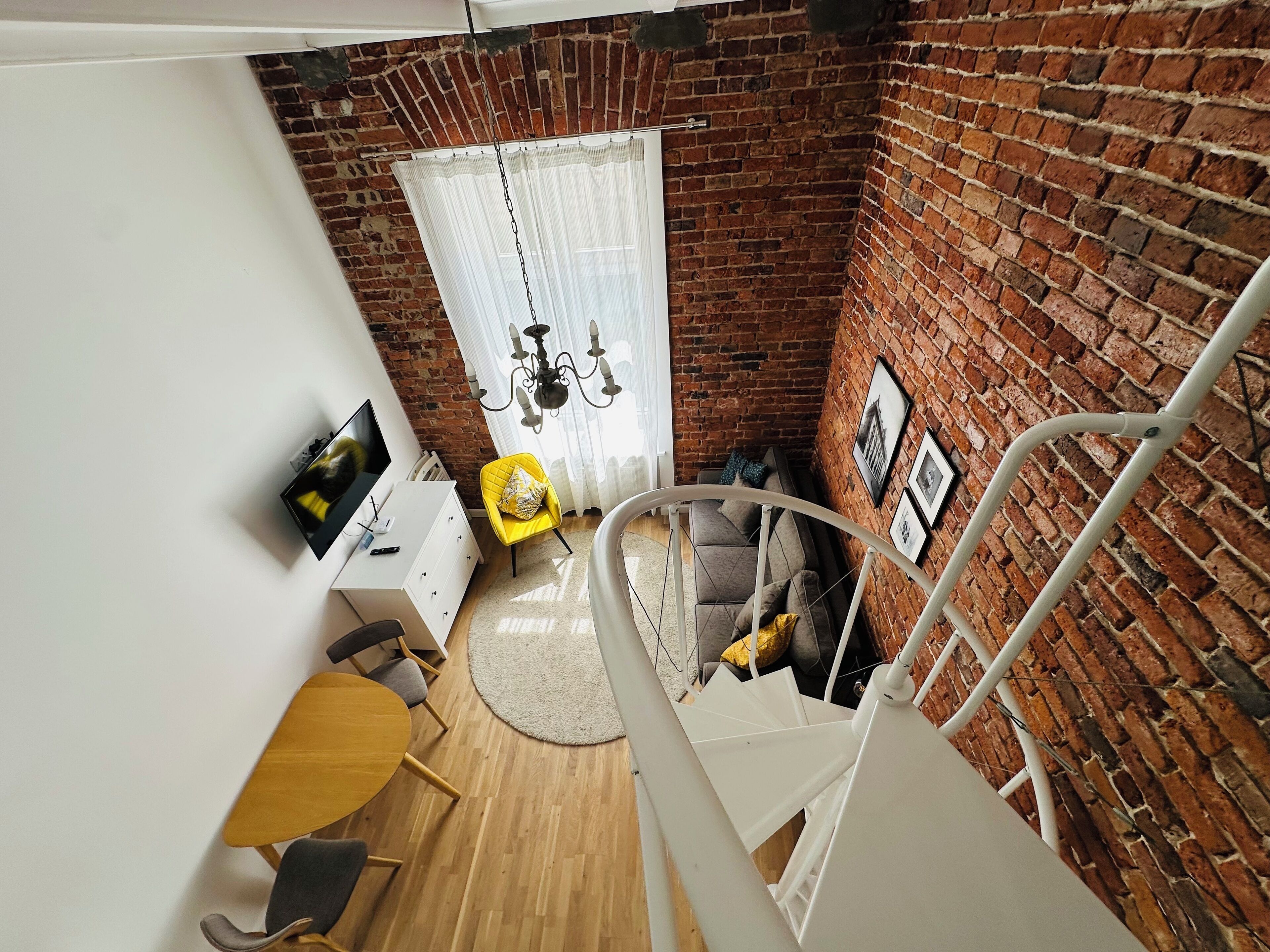 A loft-style room with exposed brick walls, a white staircase, a dining area with a round table, and a sofa.