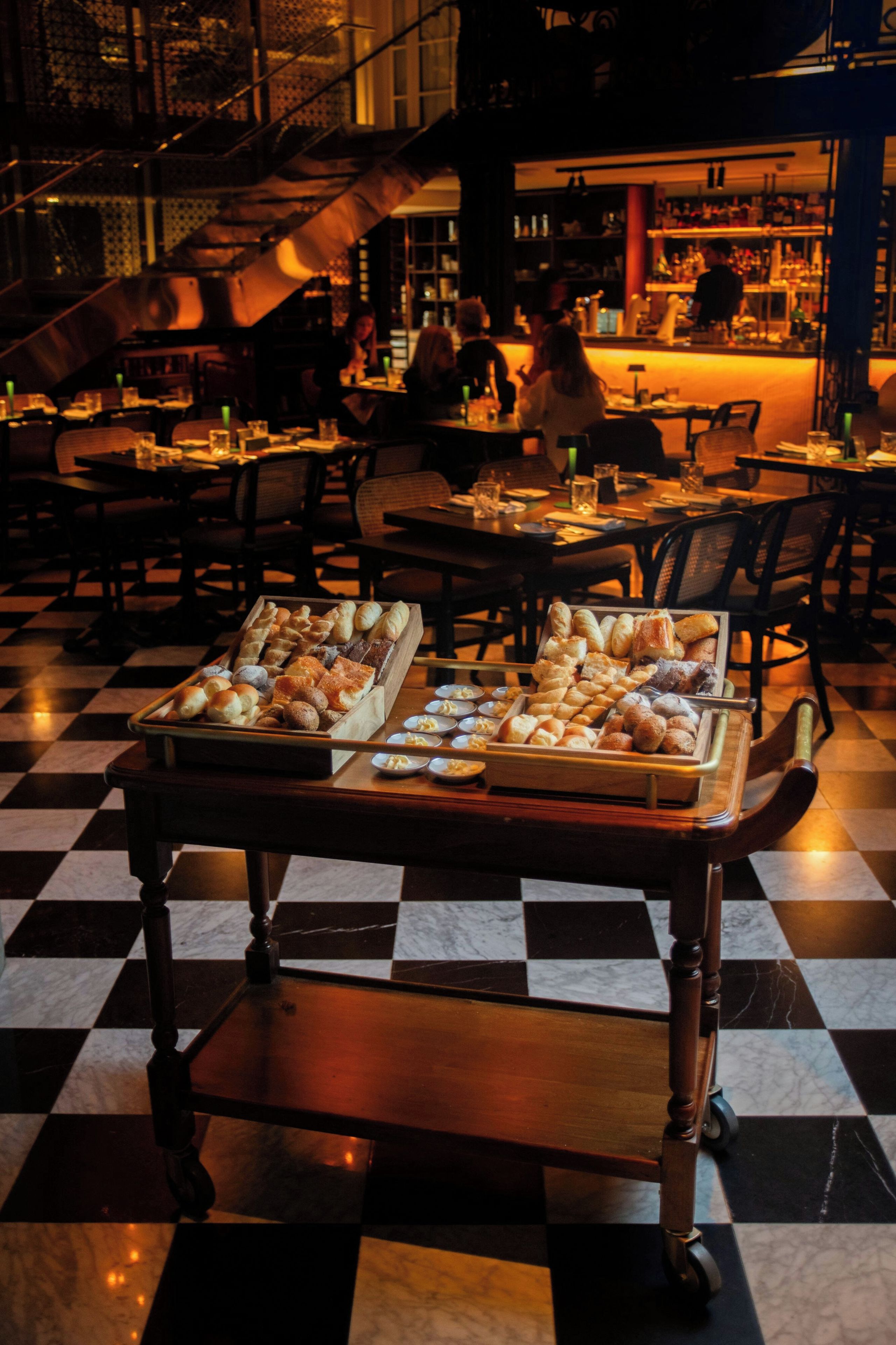 Interior de un restaurante con suelo a cuadros, una mesa de buffet con varios productos de repostería y una barra de bar al fondo.