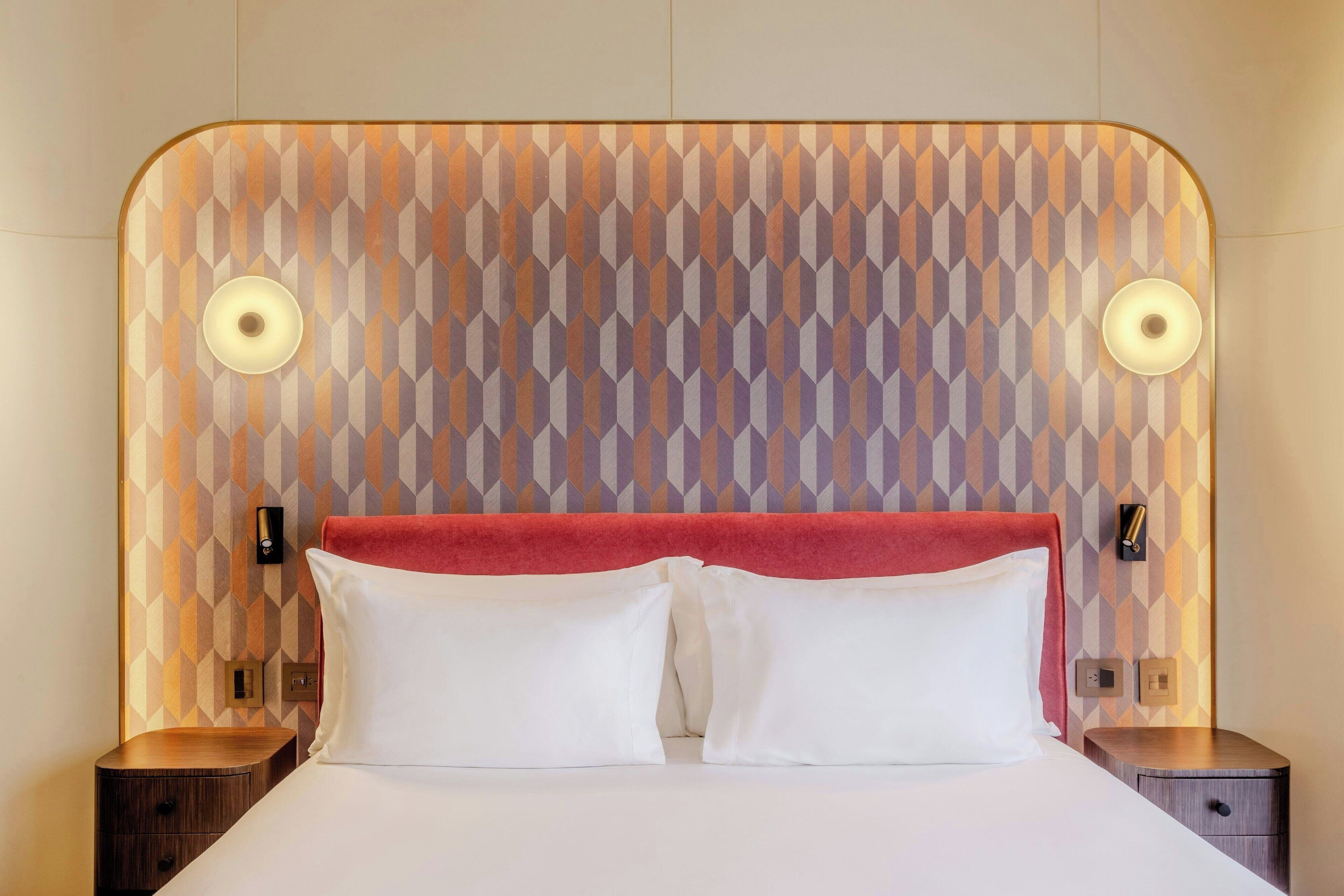 Una habitación de hotel con un cabecero de estampado geométrico, dos lámparas de noche, un detalle rojo en el cabecero y una cama bien tendida con sábanas blancas.