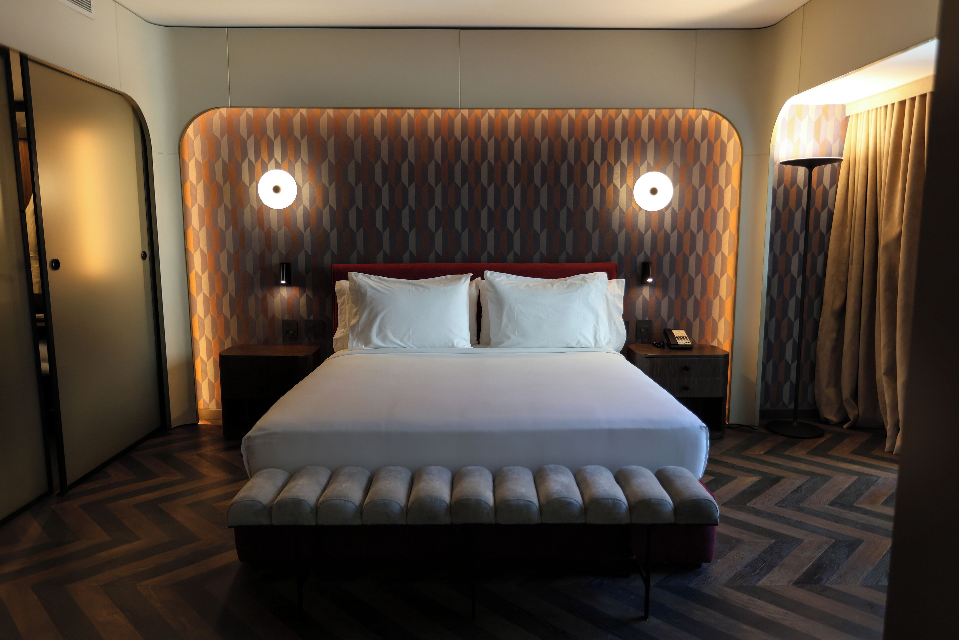 Quarto de hotel moderno com uma cama grande, duas mesas de cabeceira e cabeceira estampada.