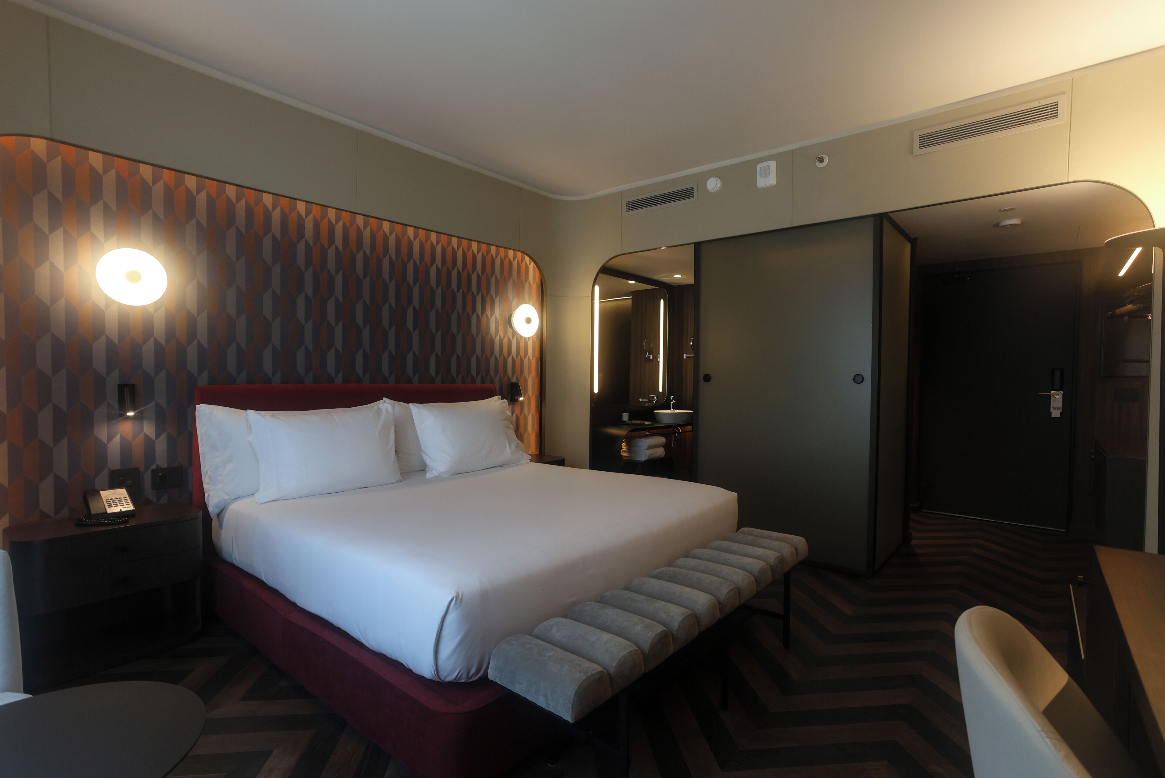 Une chambre d’hôtel avec un grand lit, une table de chevet avec une lampe, un banc au pied du lit et une salle de bain visible à travers une porte ouverte.