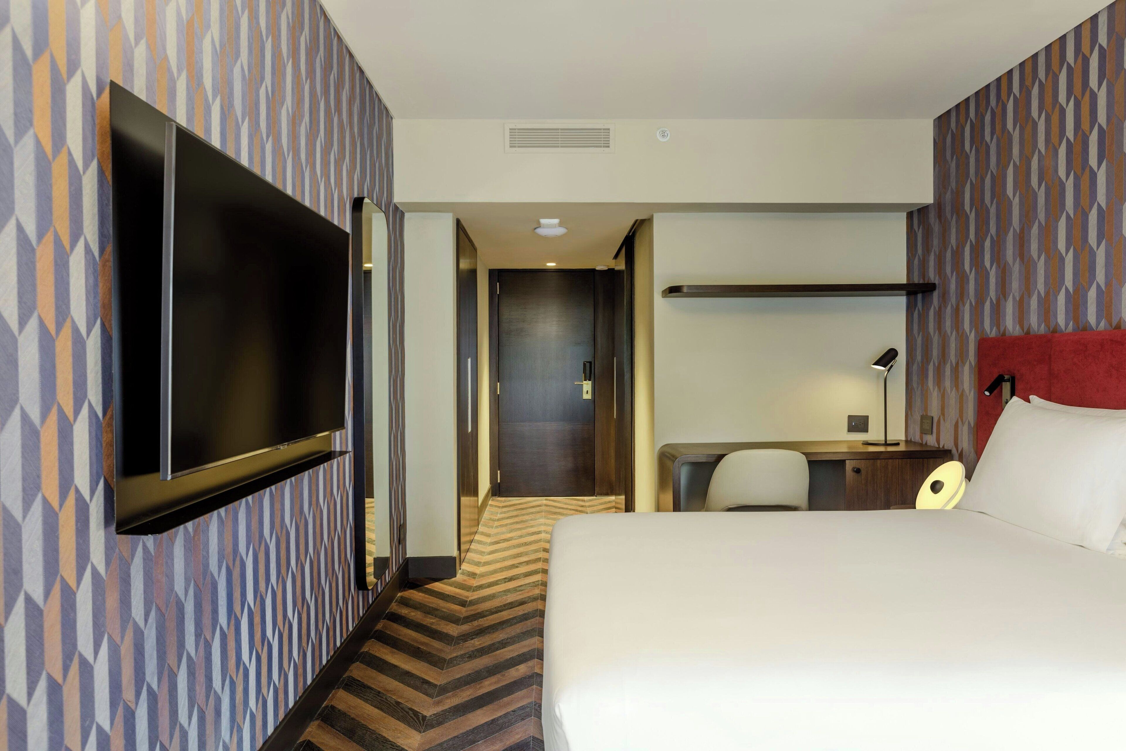 A hotel room with a large bed, a flat-screen TV mounted on the wall, a desk with a chair, and a red accent wall.