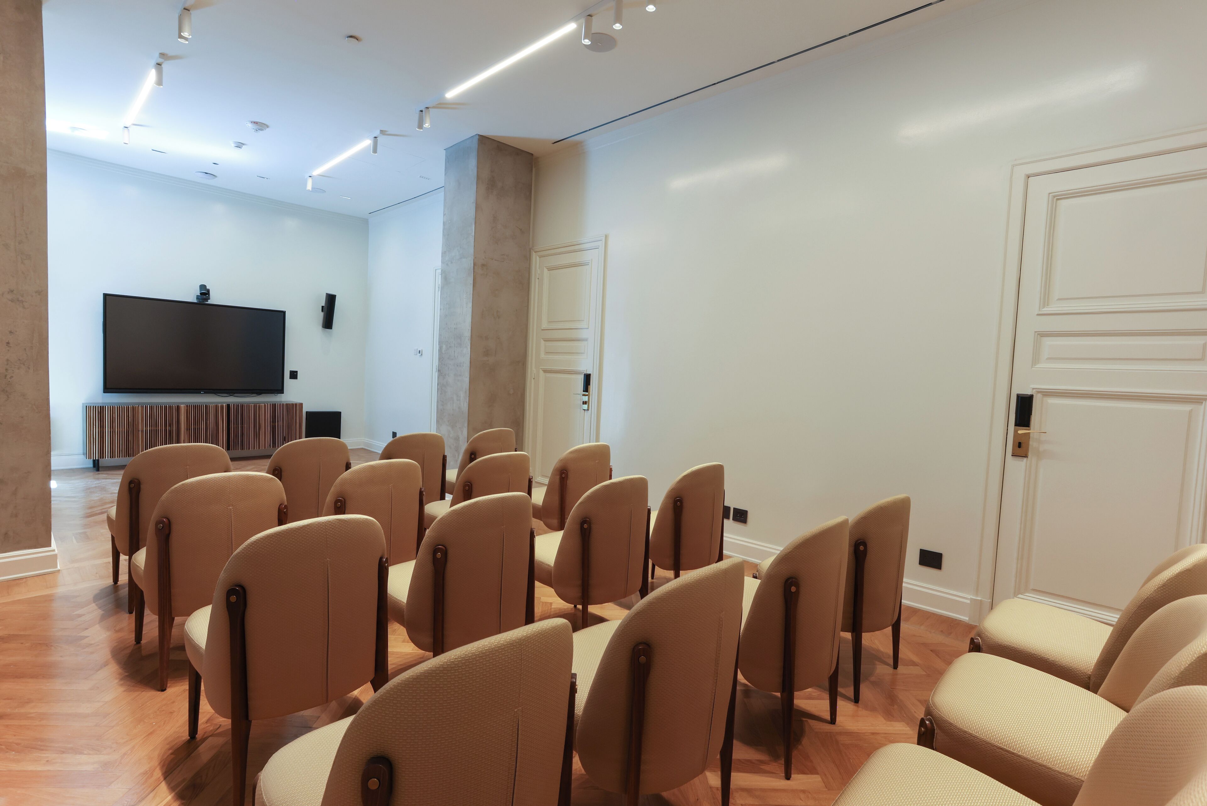 Un salón de conferencias con sillas color beige dispuestas en filas, un televisor de pantalla plana sobre un soporte y suelo de madera.