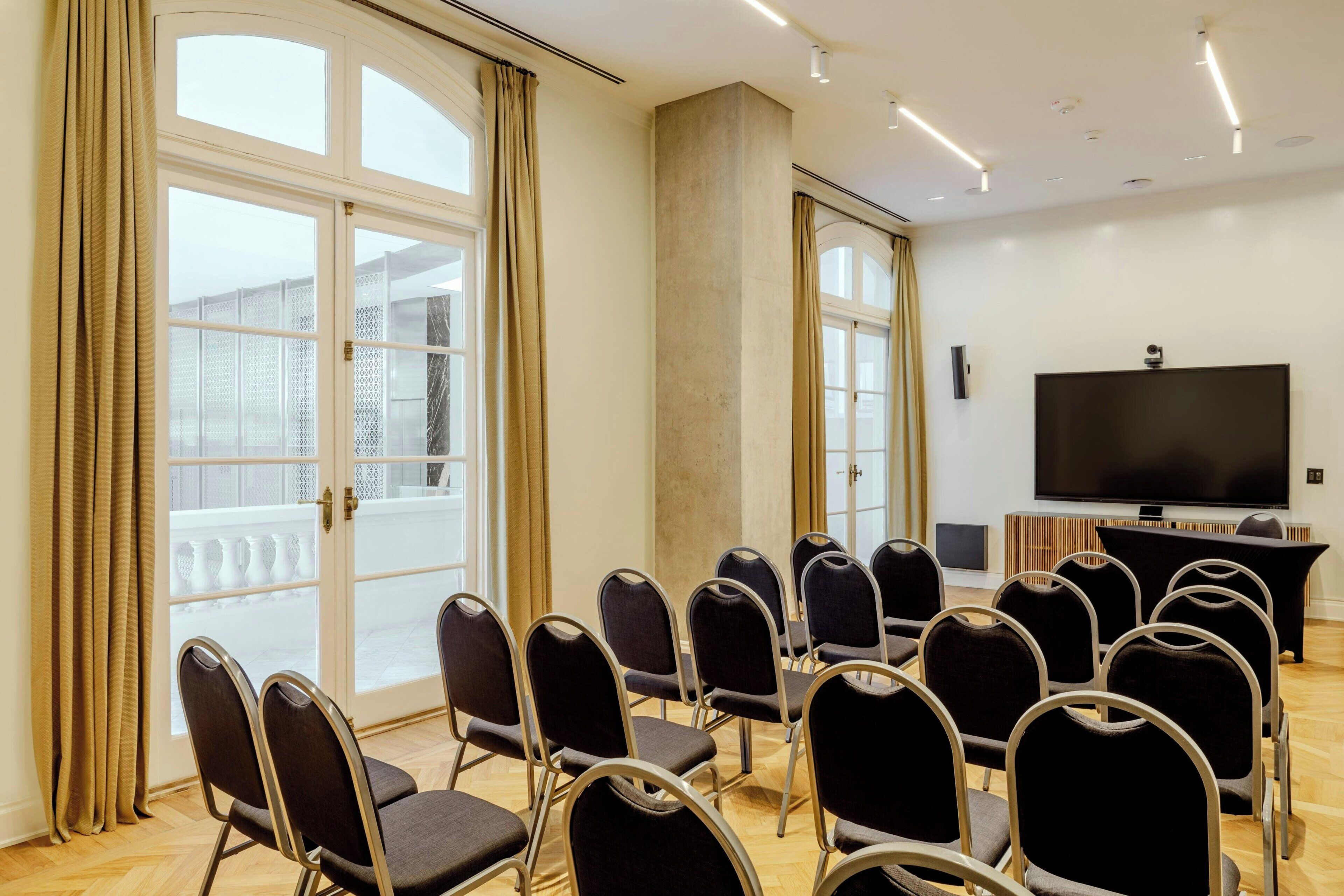 Ein Konferenzraum mit einem großen Fernseher, mehreren Stühlen und großen Fenstern mit Vorhängen.