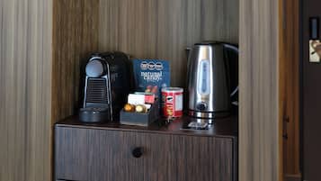 Una pared de madera con un estante que sostiene dos tazas blancas y una tetera. Abajo, hay un cajón con una caja de caramelos, una lata de refresco y una cafetera.