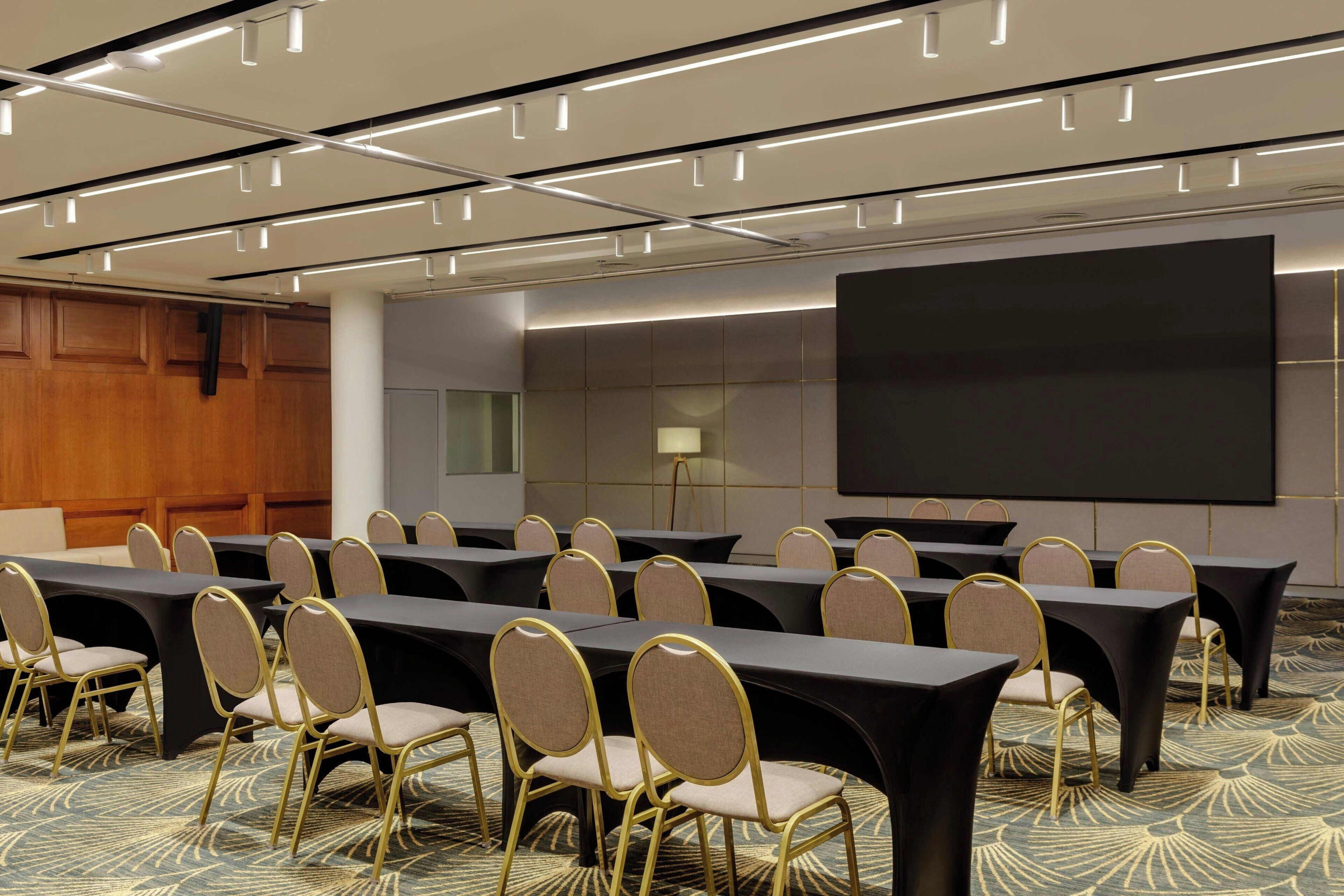 Un salón de conferencias con varias mesas y sillas, una pantalla grande y piso alfombrado.