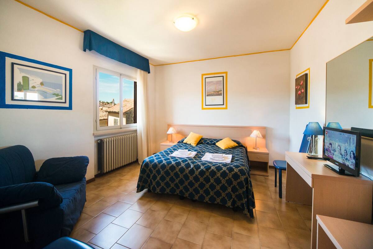 Habitación de hotel con cama, escritorio, silla, ventana con vistas y cuadros en las paredes.