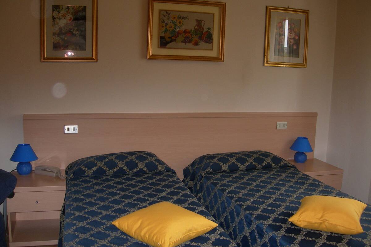 Habitación de hotel con dos camas, una mesita de noche, una lámpara y tres cuadros en la pared.