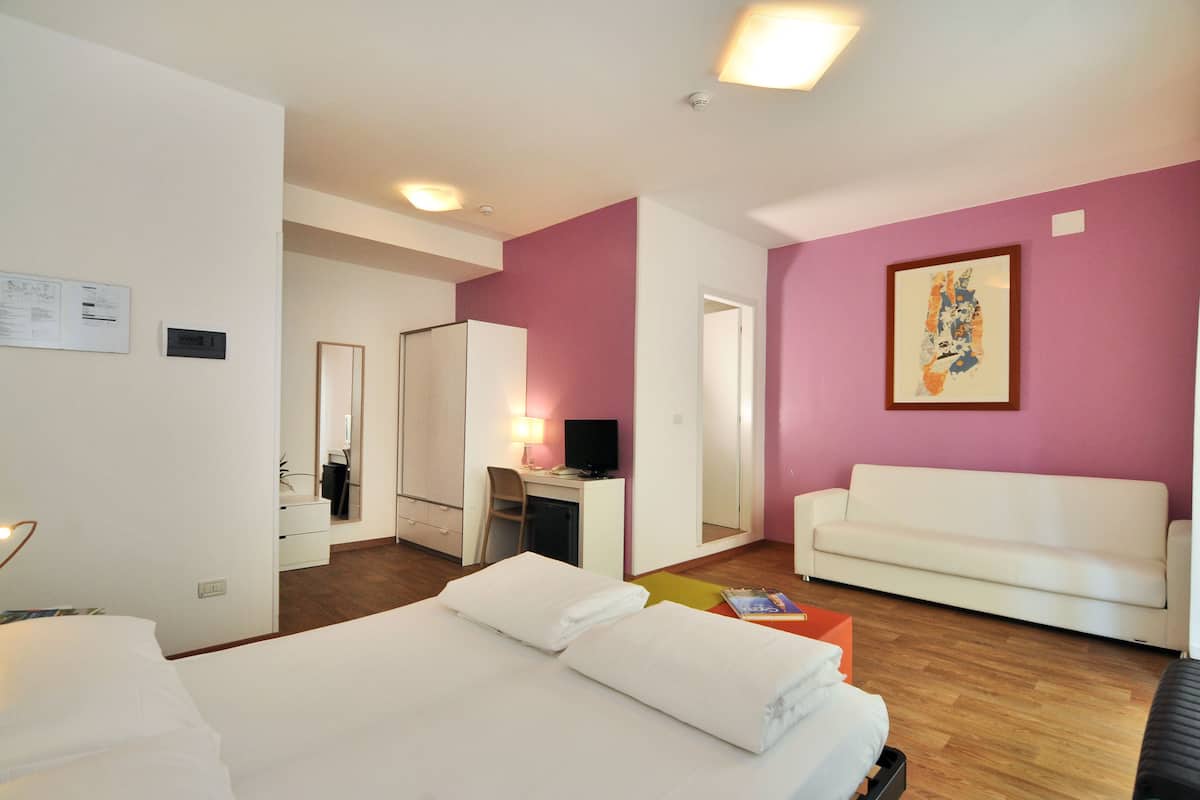 A hotel room with a bed, a desk, a chair, a white sofa, and a painting on the wall.