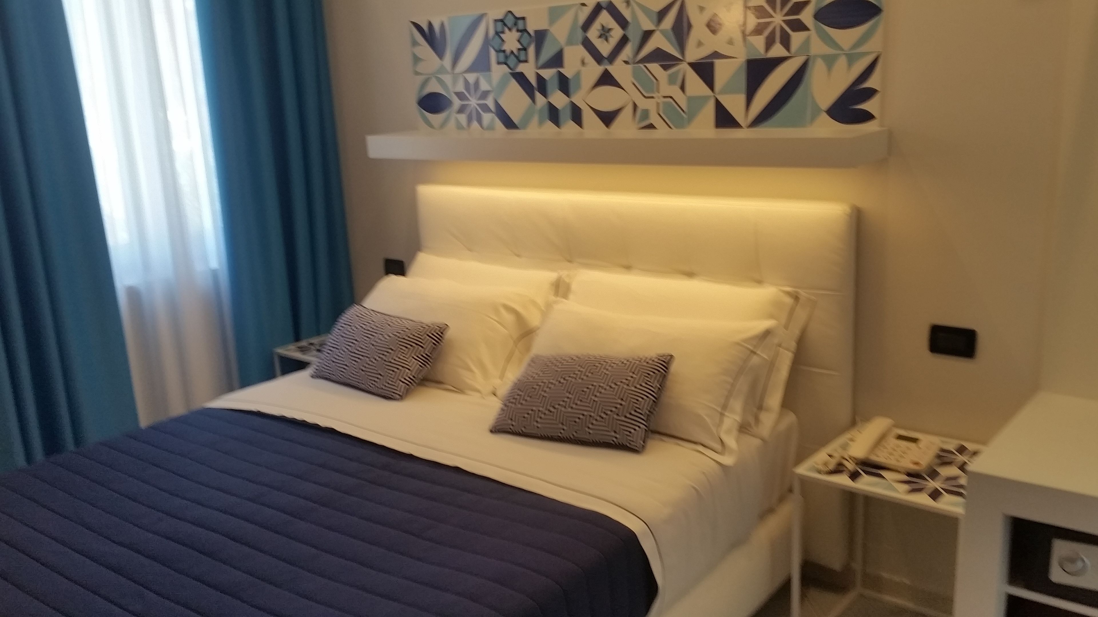 A modern bedroom with a white bed, two patterned pillows, a dark blue bedspread, a geometric wall decoration, and a nightstand with a phone and a small table.