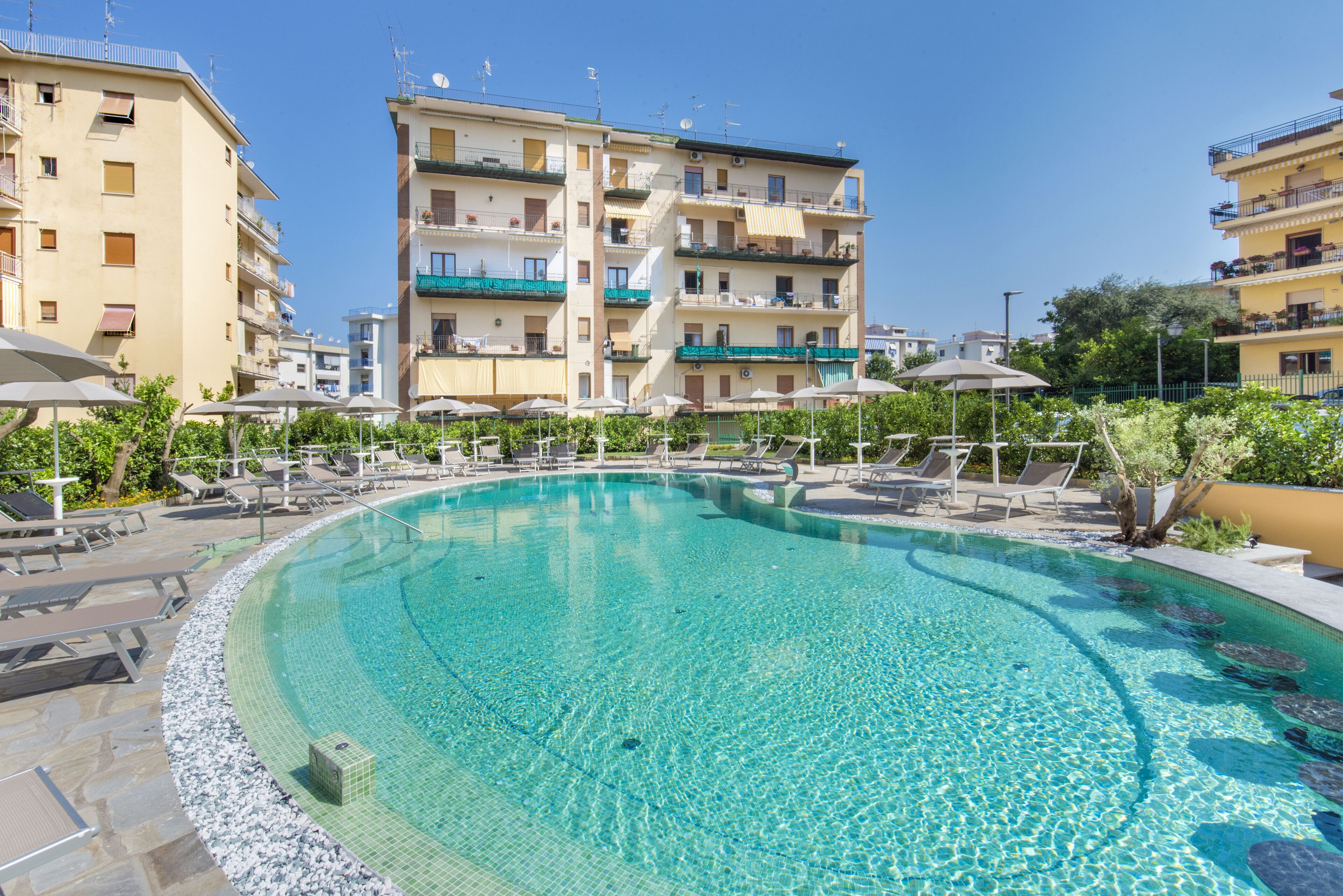 A swimming pool with lounge chairs and umbrellas, surrounded by multi-story buildings with balconies.