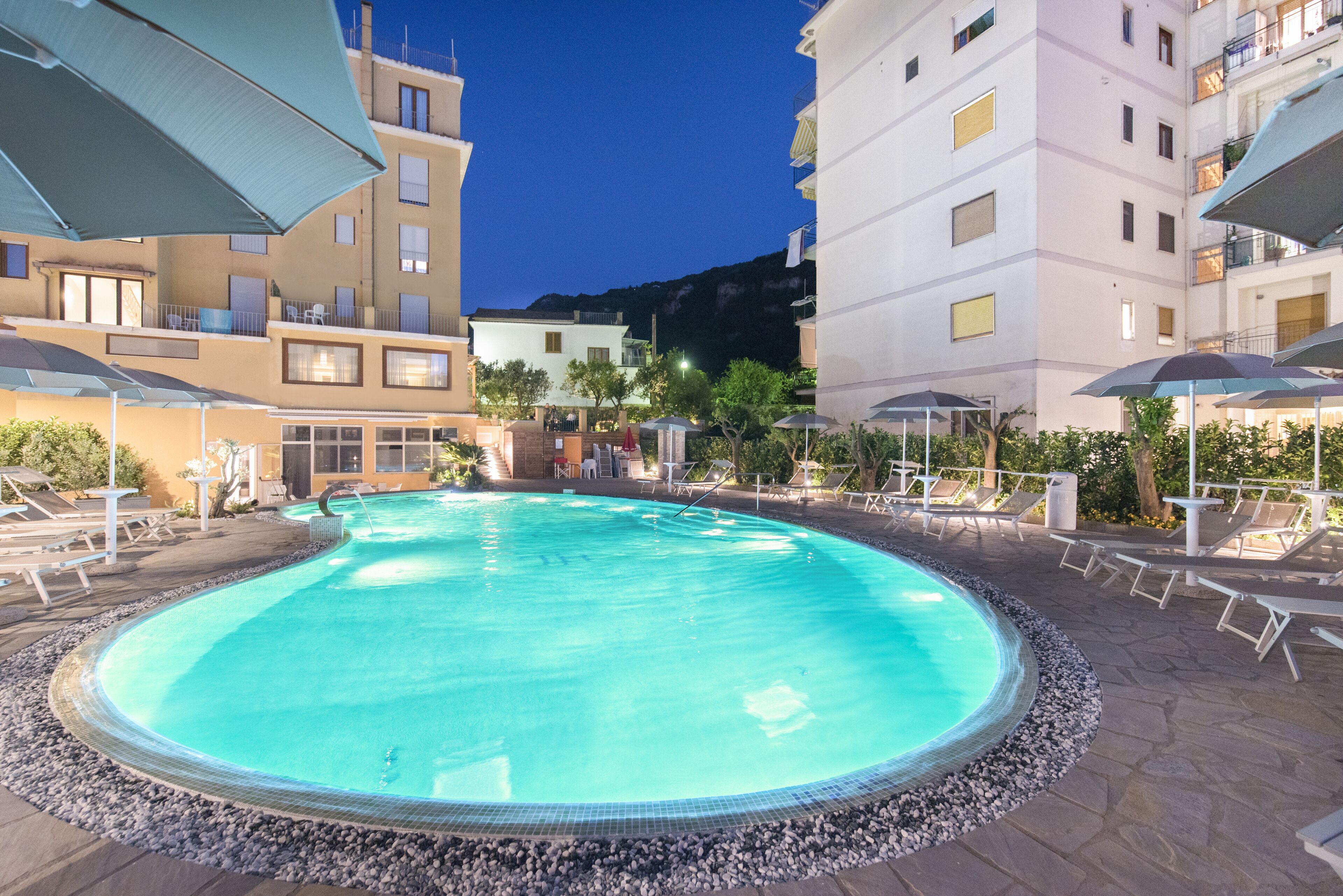 A circular swimming pool with lounge chairs and umbrellas, surrounded by buildings with balconies and outdoor seating.