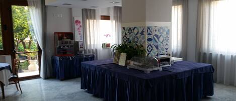 Une salle de conférence avec une grande table recouverte d’une nappe bleue, entourée de chaises. On y trouve un distributeur automatique et une petite plante sur la table. La salle a un sol carrelé et de grandes fenêtres avec des rideaux.