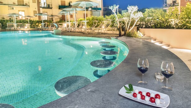 A pool area with lounge chairs and umbrellas, a table with glasses and a plate of fruit, and a building in the background. - Hotel Leone (Sorrento)