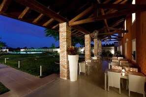 An outdoor dining area with brick pillars, white tables, and a pool in the background. - Antico Borgo Torricella (San Vito al Tagliamento)