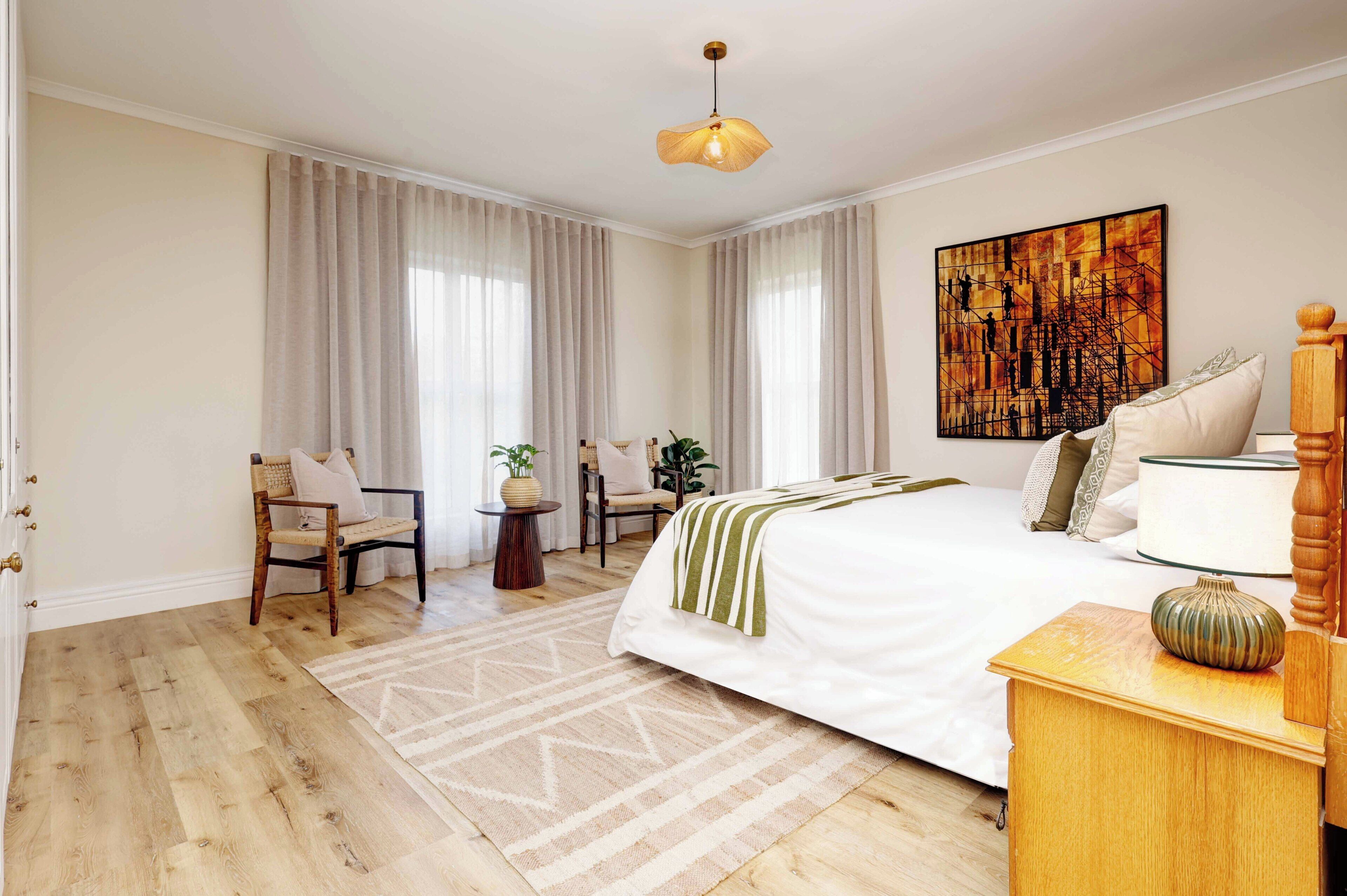 A bedroom with a large bed, a bedside table, a chair, a small table with a plant, and a painting on the wall.