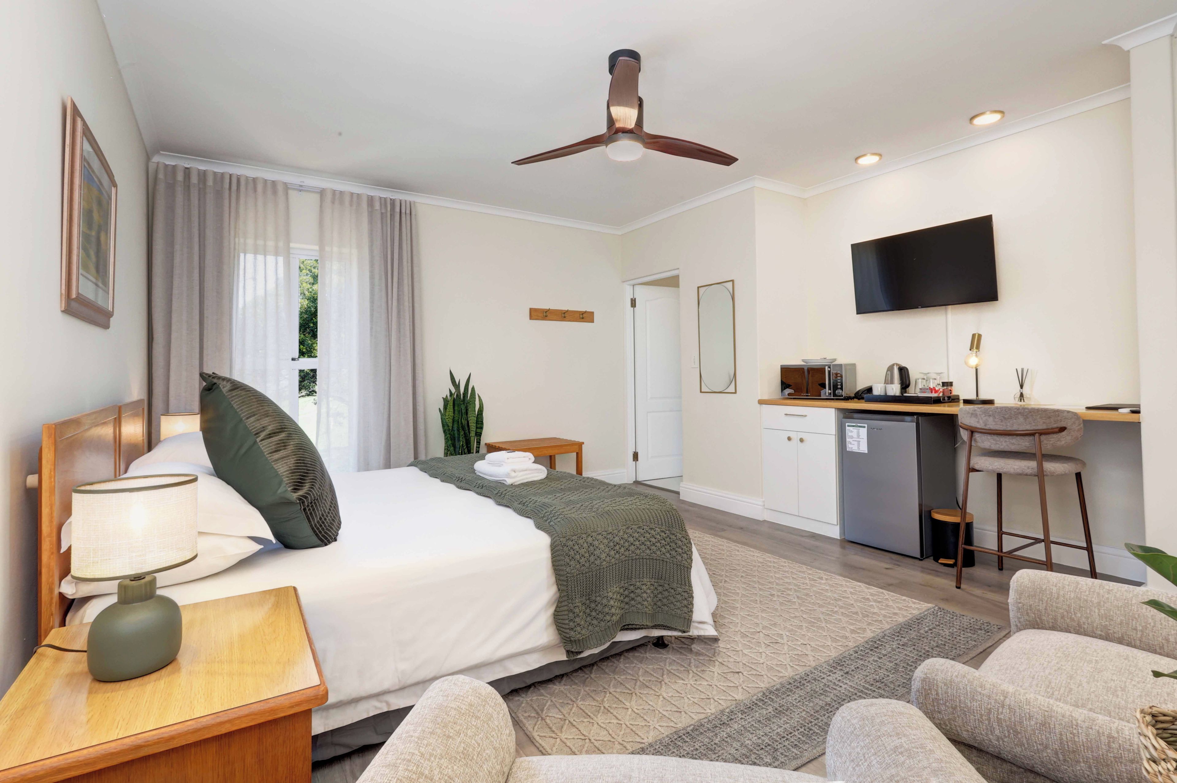 A hotel room with a bed, bedside table, lamp, curtains, a ceiling fan, a TV, a small kitchenette, and a seating area with a chair and a sofa.