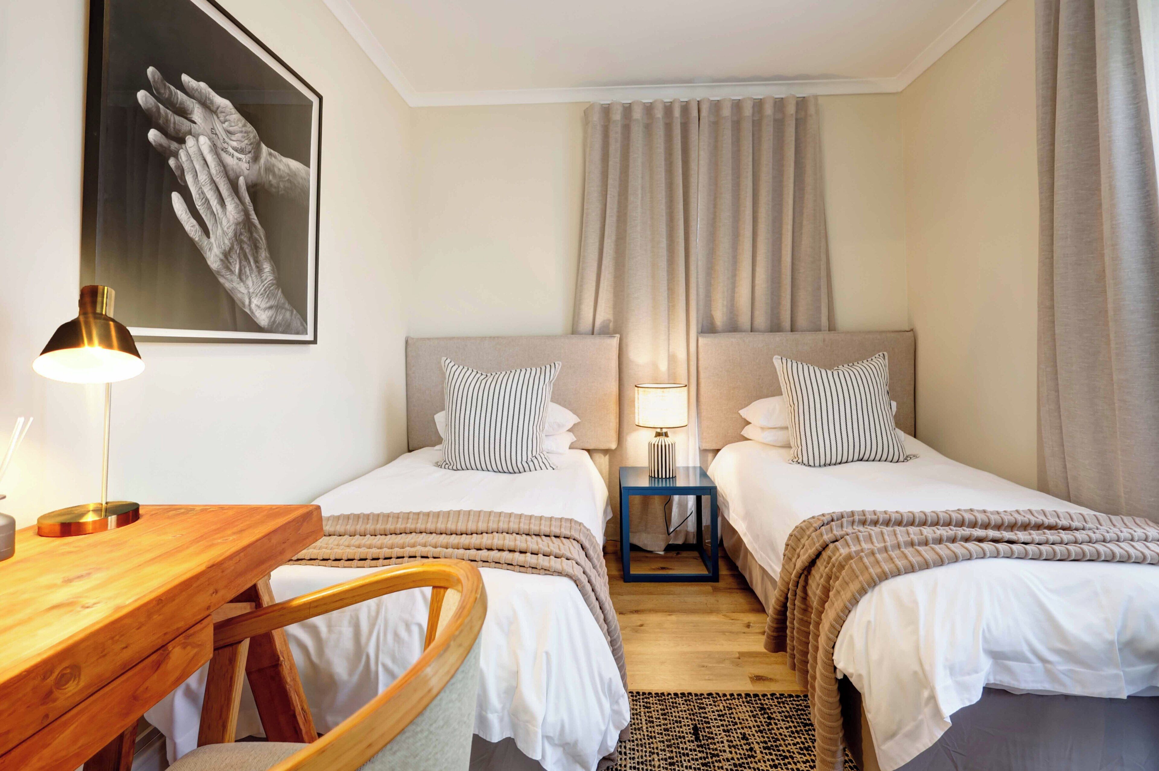 A hotel room with two beds, a desk, a chair, a lamp, and a painting on the wall.
