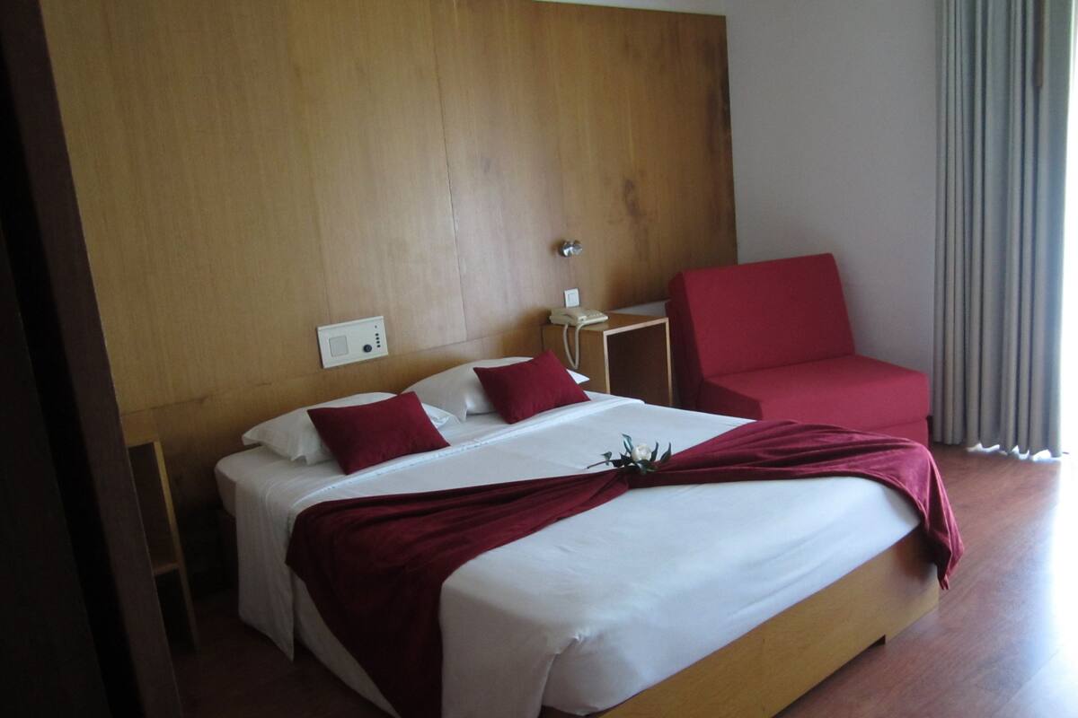 A hotel room with a wooden headboard, a large bed with white linens and red pillows, a red armchair, a bedside table with a phone, and a window with curtains.