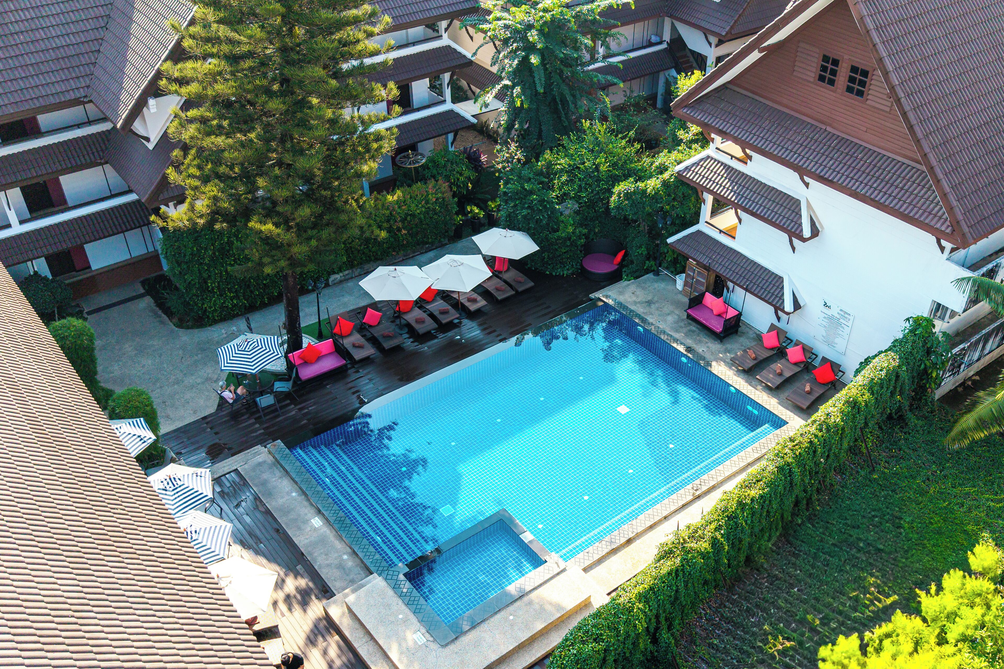 An aerial view of a hotel with a swimming pool surrounded by lounge chairs and umbrellas, a covered seating area, and lush greenery.