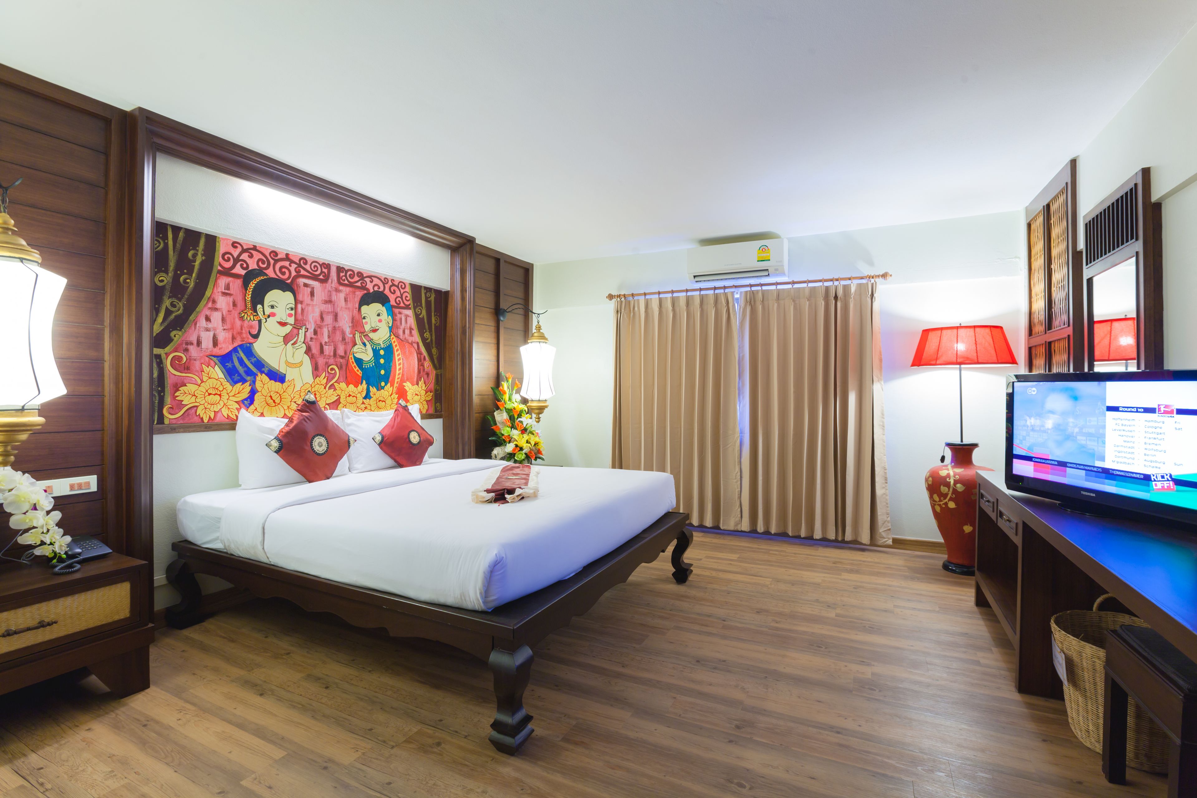 A hotel room with a bed, a television, a desk, a lamp, a vase, and a painting on the wall.