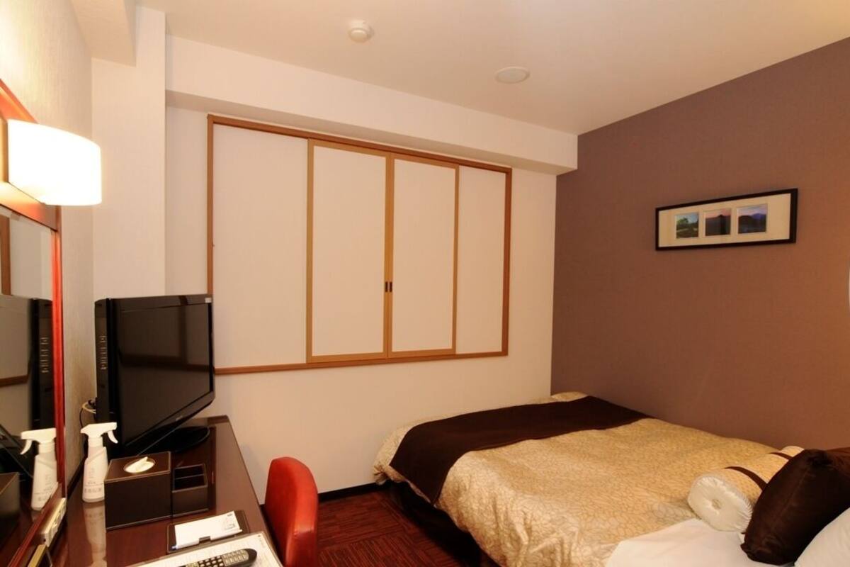 A hotel room with a bed, a desk with a television, a mirror, and a picture on the wall.