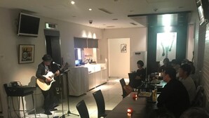 Un área de comedor informal con un músico actuando en un pequeño escenario, mesas con comensales y una cocina al fondo.