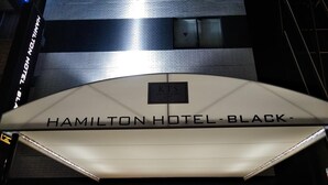 La entrada al Hotel Hamilton, de color negro, presenta un diseño moderno con un cartel sobre la entrada y un camino bien iluminado que conduce al interior.
