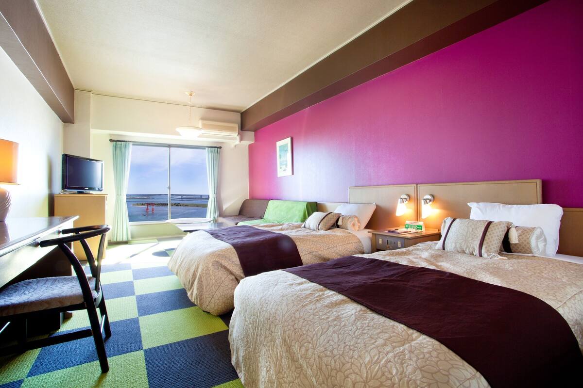 Ein Hotelzimmer mit zwei Betten, einem Schreibtisch, einem Stuhl, einem Fernseher und einem Fenster mit Aussicht.