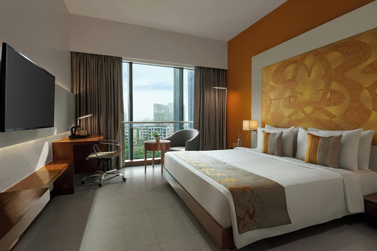 A hotel room with a large bed, a desk, a chair, a TV, and a view of the city.