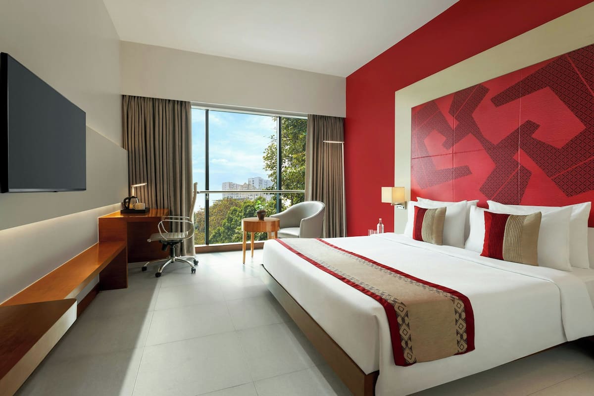 A hotel room with a large bed, a desk, a chair, a TV, and a window with a view of buildings and trees.