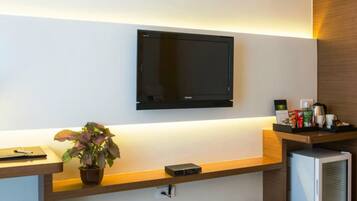 A hotel reception area with a flat-screen TV mounted on the wall, a wooden reception desk, a potted plant, a coffee machine, and a refrigerator.