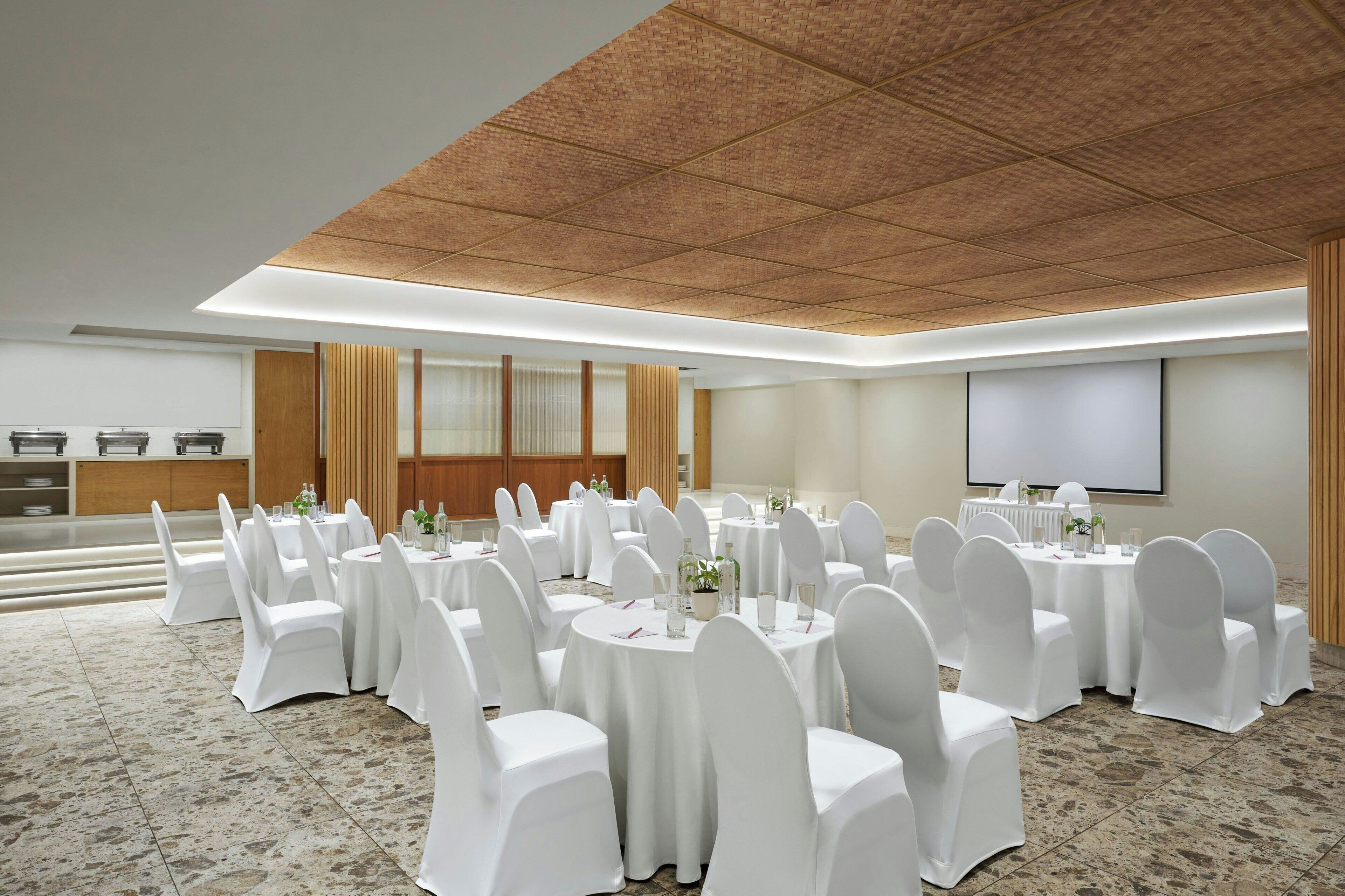 A conference room with round tables, white chairs, a projector screen, and a wooden ceiling.